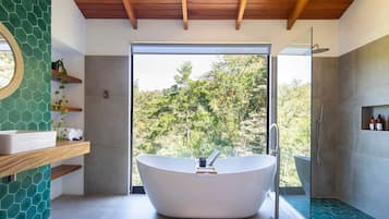 A modern bathroom with a freestanding bathtub, a glass-enclosed shower, and a view of trees through a large window.