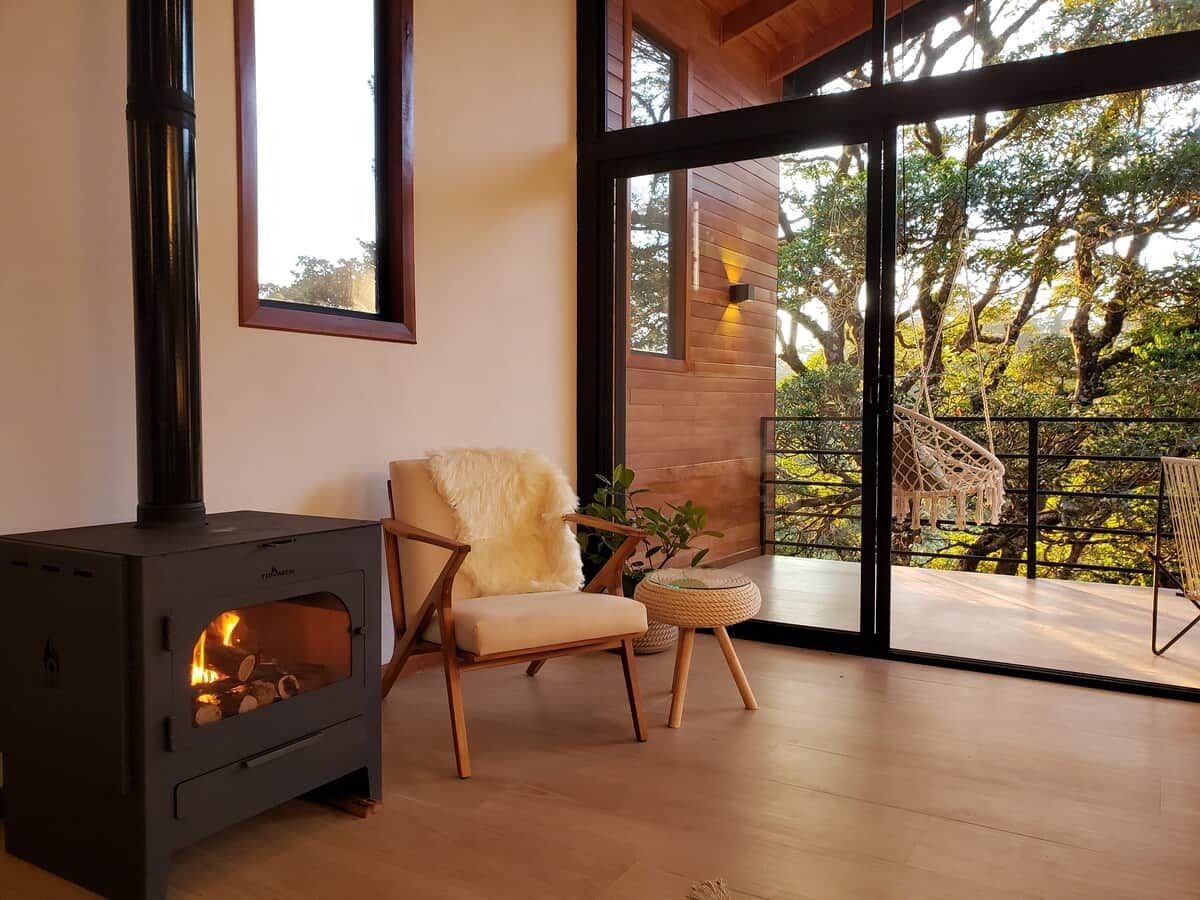 Een modern interieur met een houtkachel, een comfortabele fauteuil en uitzicht door grote glazen deuren op een terras.