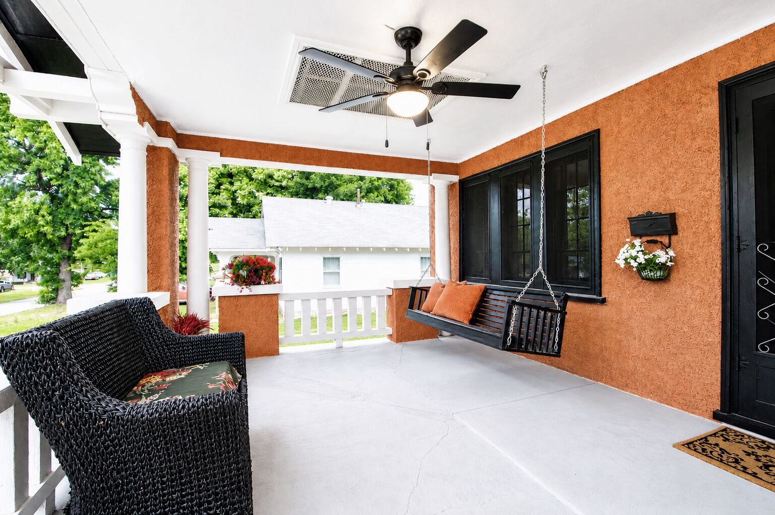 A covered porch with a swing, a wicker chair, a ceiling fan, and a door with a decorative handle.