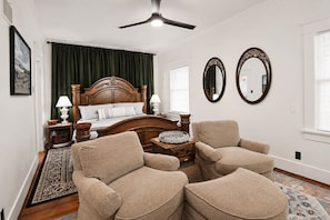 A bedroom with a wooden bed, two armchairs, a nightstand, a lamp, and a mirror.