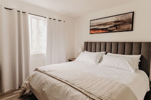 Een slaapkamer met een groot bed, een hoofdeinde, een nachtkastje, een lamp en een raam met gordijnen.