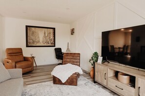 Een woonkamer met een bank, een bruine fauteuil, een salontafel, een televisie en een schilderij aan de muur.