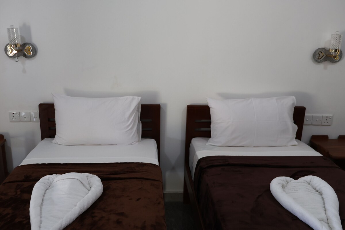Habitación de hotel con dos camas, cada una con un almohadón blanco y una manta marrón. Hay dos mesitas de noche con lámparas, y las paredes presentan un diseño sencillo con luminarias.