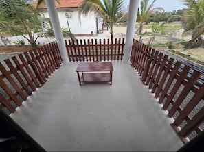 Un espacio de estar al aire libre techado con un banco y una mesa de madera, rodeado por una valla de madera, con vistas a un edificio blanco y palmeras al fondo.