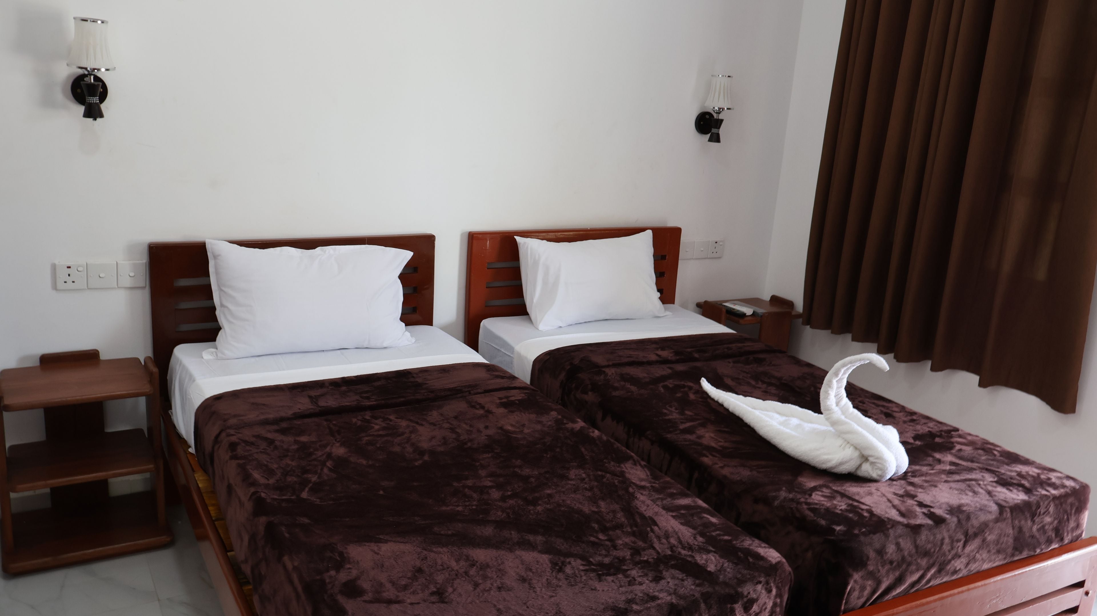 A hotel room with two beds, each with a wooden headboard, white bedding, and a brown velvet bedspread. There are two bedside tables with lamps, a brown curtain, and a wall socket.
