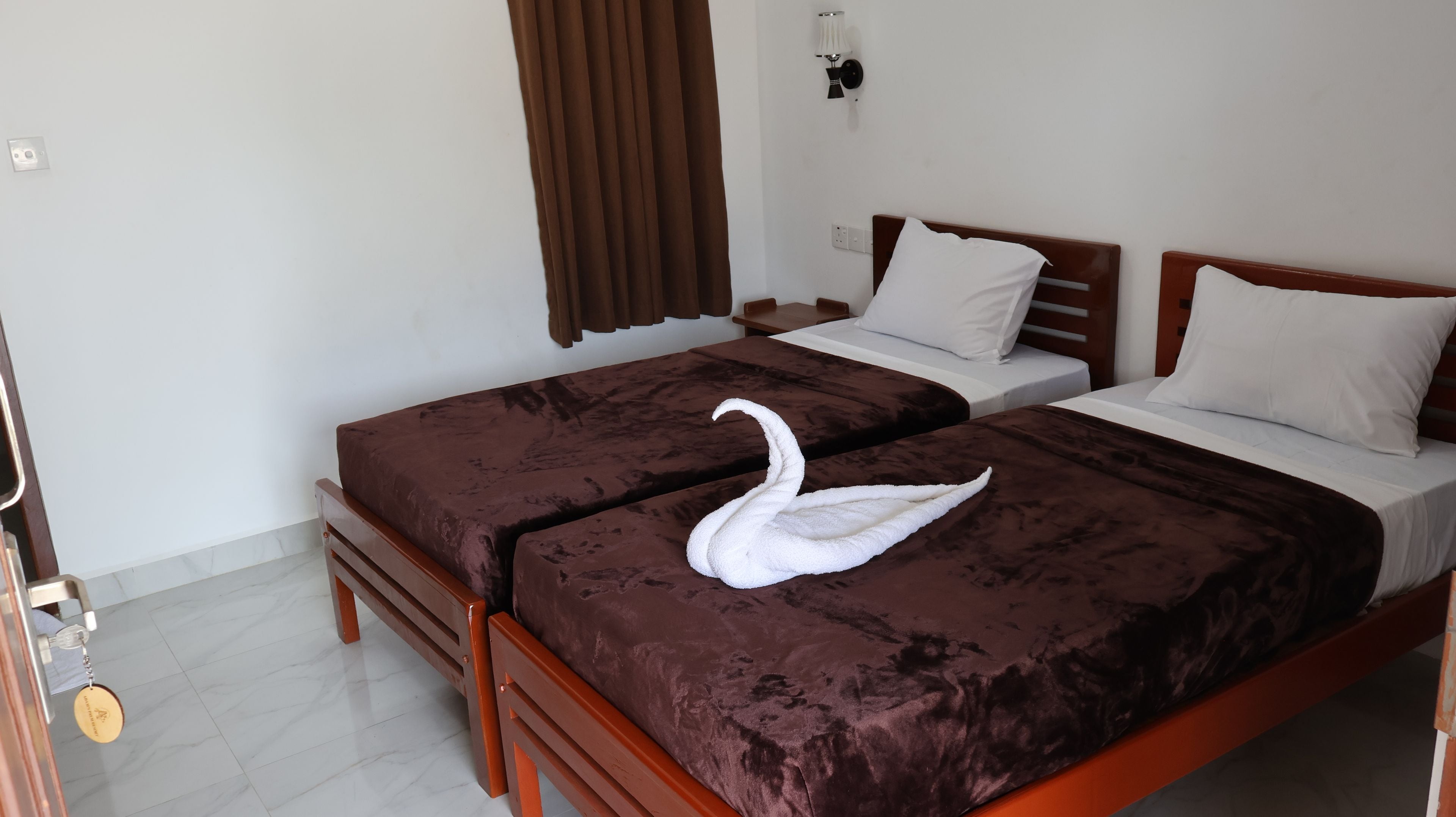 A hotel room with two beds, a brown curtain, a wall-mounted lamp, and a keychain on the door.