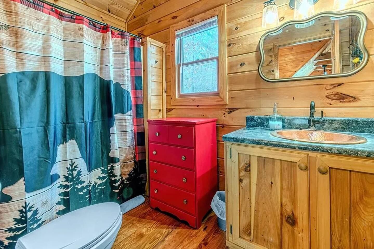 A wooden cabin bathroom with a toilet, a red dresser, a sink with a granite countertop, and a window.