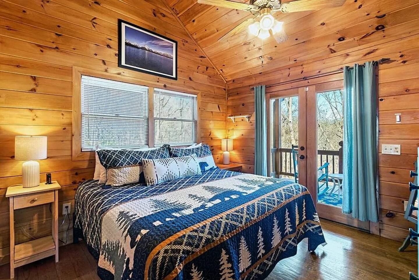 A wooden cabin room with a bed, a nightstand, a window with blinds, a ceiling fan, and a view of the outdoors.