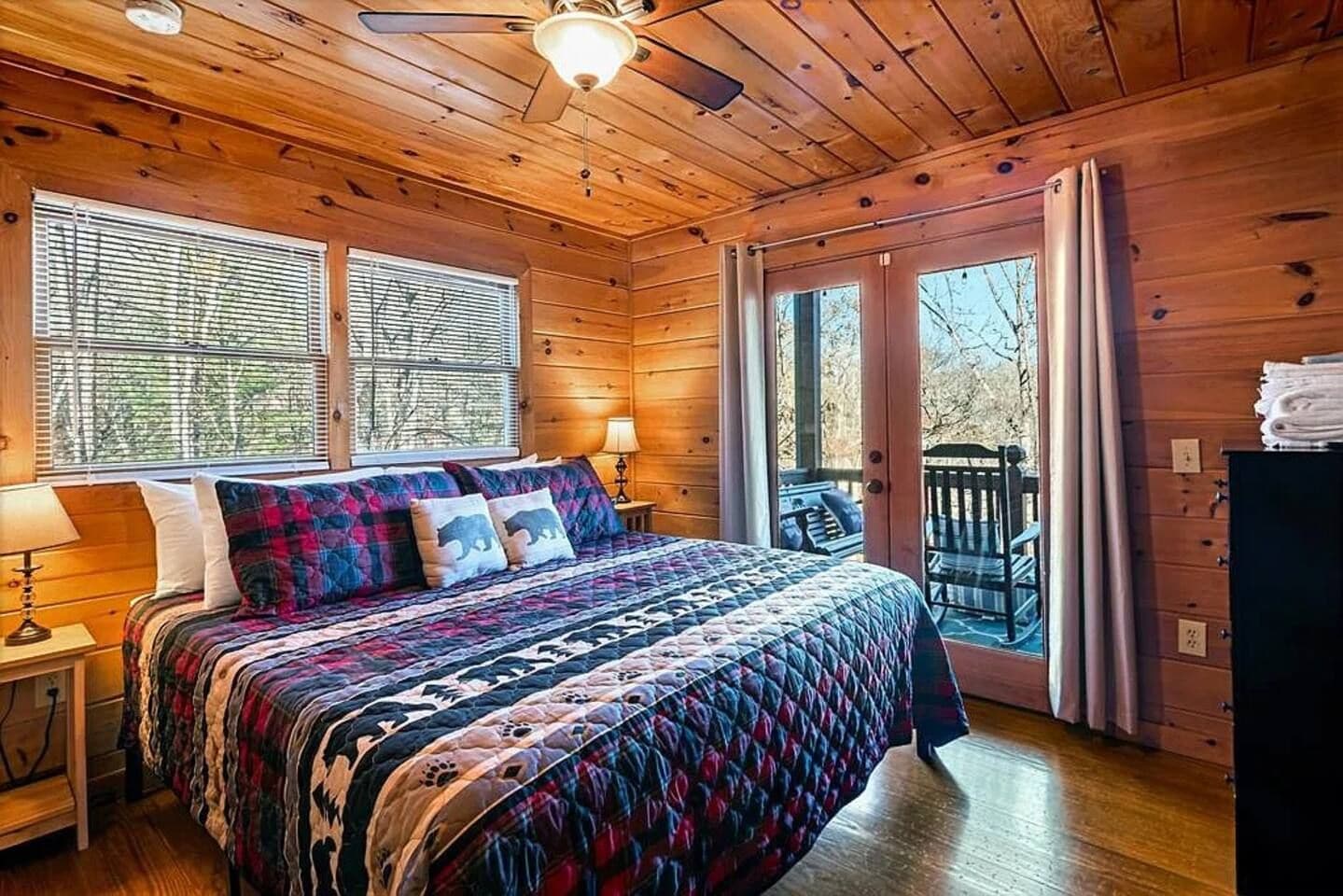 Un dormitorio tipo cabaña de madera con cama, mesita de noche, ventana con persianas y puerta que da a una terraza al aire libre.