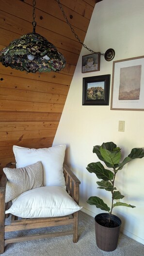 A cozy room with a wooden ceiling, a hanging mosaic light fixture, a wooden chair with cushions, a potted plant, and framed artwork on the wall. - The Collected Cottage (Cambria)