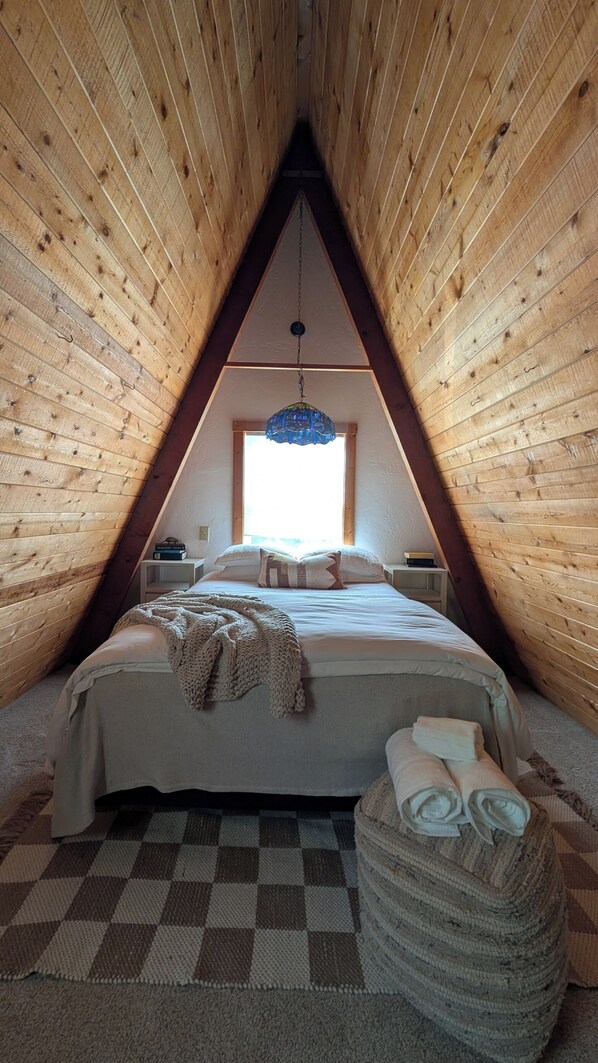 A cozy attic bedroom with a sloped ceiling, a large bed, a hanging light fixture, and a window providing natural light. - The Collected Cottage (Cambria)