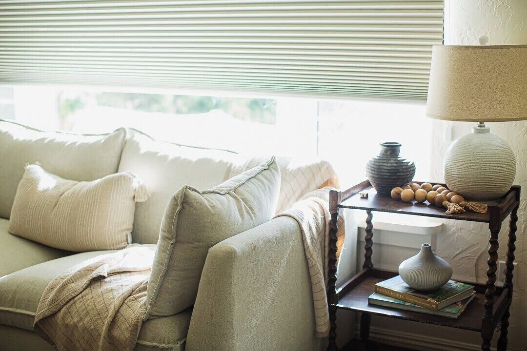 A cozy living room with a sofa, a side table with a lamp, and a window with blinds.