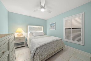 Een slaapkamer met een bed, nachtkastje, dressoir, plafondventilator, raam met jaloezieën en een wanddecoratie.