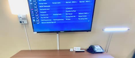A hotel room with a television displaying a schedule, a desk with a telephone, and a built-in mini-fridge.