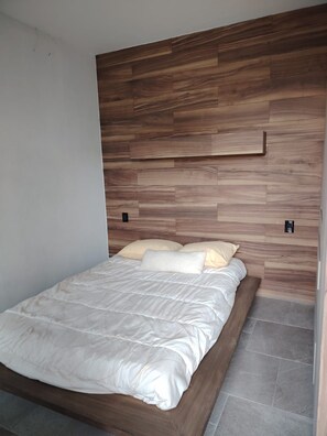 A bed with a wooden headboard and a white mattress in a room with grey tiled flooring.