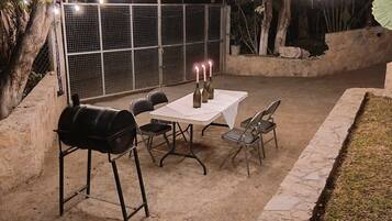 Patio esterno con barbecue, tavolo, sedie e luci decorative appese.