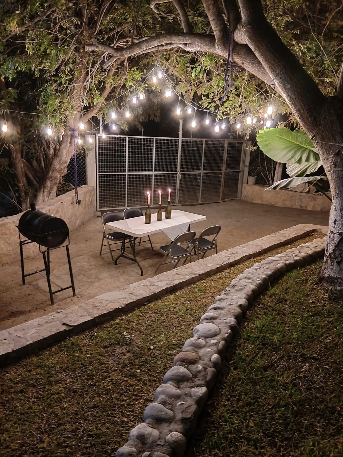 Un patio al aire libre con mesa y sillas, luces colgantes y una parrilla.