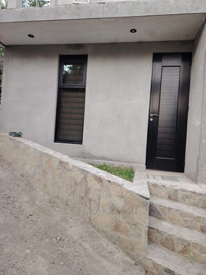 A modern building with a concrete facade, a window with horizontal blinds, a door with a vertical handle, and a stone retaining wall.
