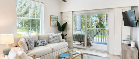 A living room with a sofa, armchairs, a coffee table, and a TV. There is a balcony with a chair and a table.