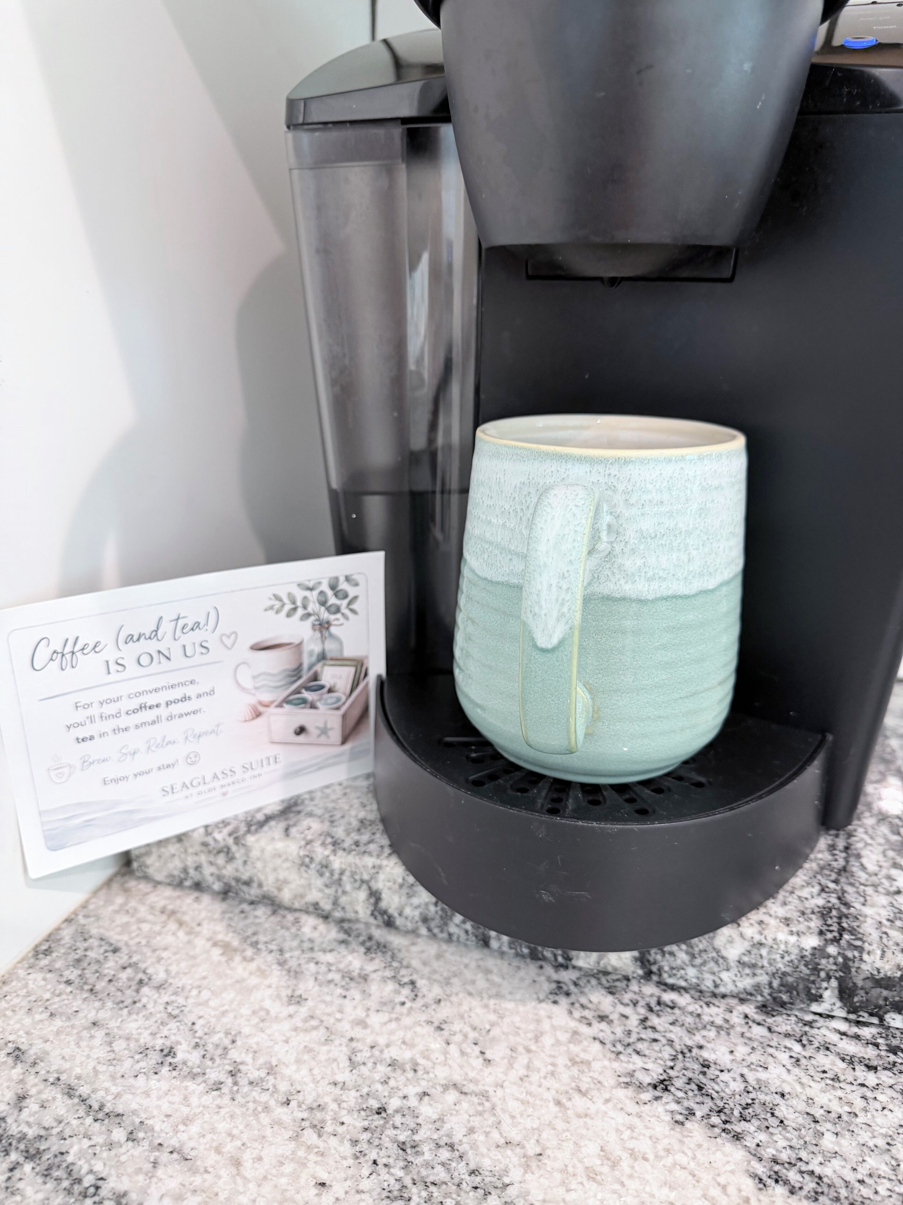 Une machine à café avec une tasse en céramique posée sur le bac d’égouttement, un panneau indiquant les types de café et de thé proposés, et un comptoir en granit.