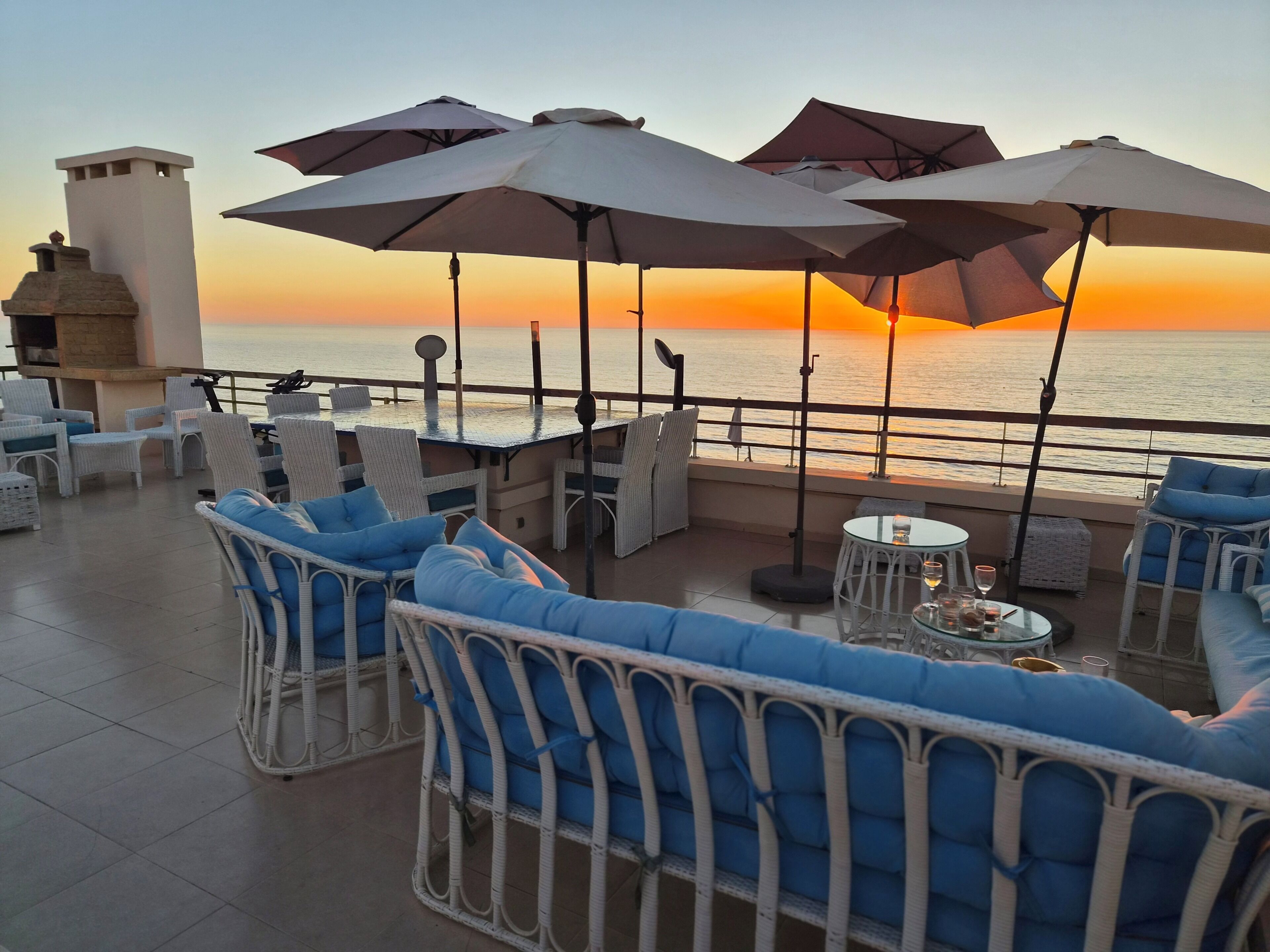 Une terrasse dotée de meubles en osier, de parasols et offrant une vue sur la mer au coucher du soleil.