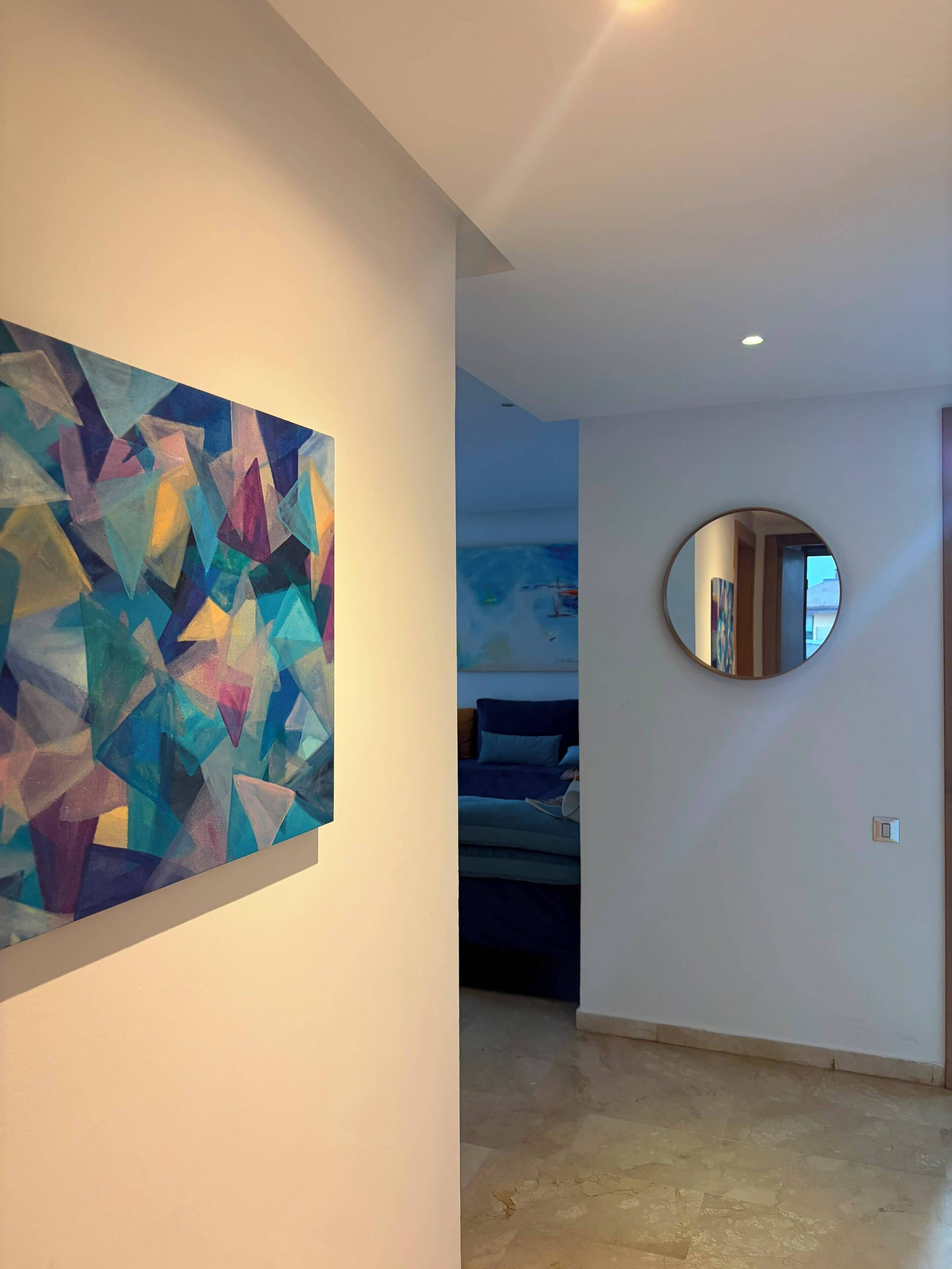 A hallway with a geometric abstract painting on the left, a round mirror on the right, and a glimpse of a bed in the background.