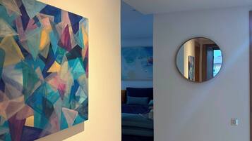 A hallway with a geometric abstract painting on the left, a round mirror on the right, and a glimpse of a bed in the background.