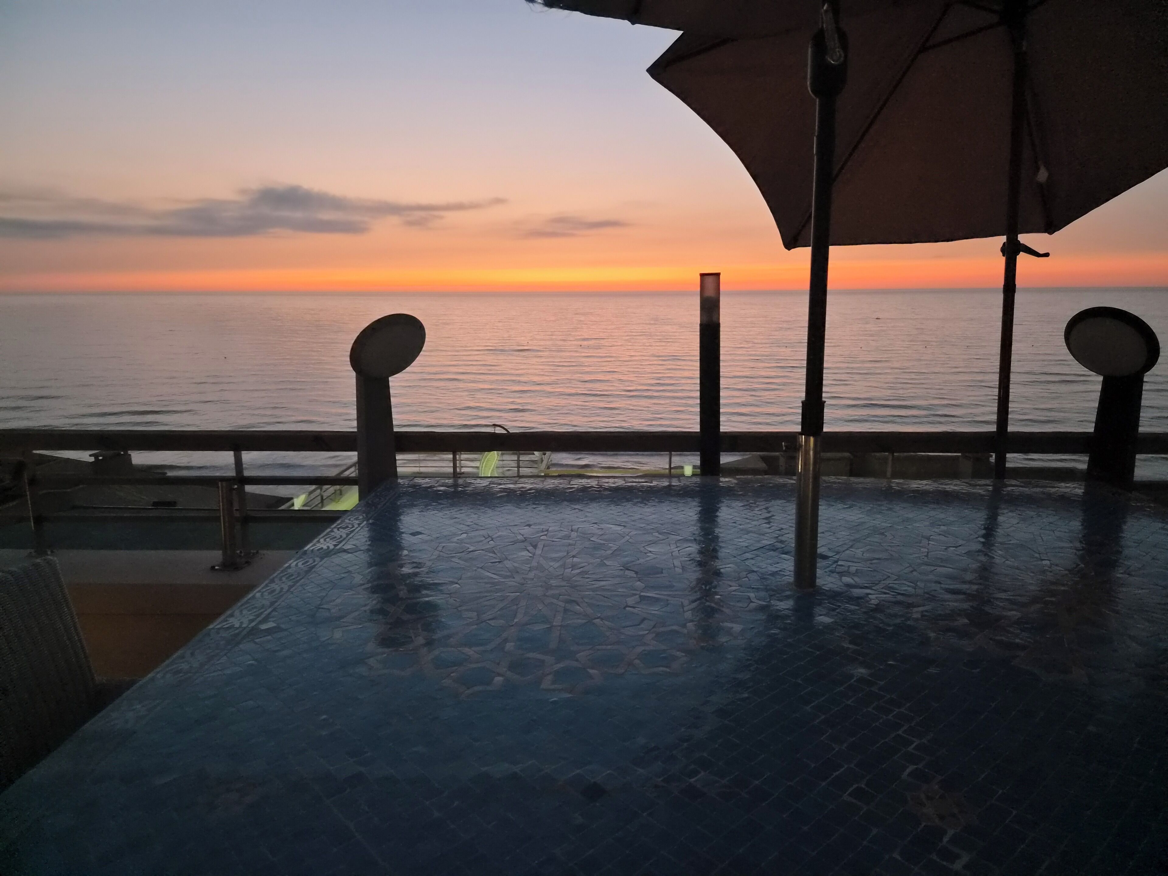 Una terraza con vistas a la puesta de sol sobre el agua, con suelo de baldosas y barandillas de metal.