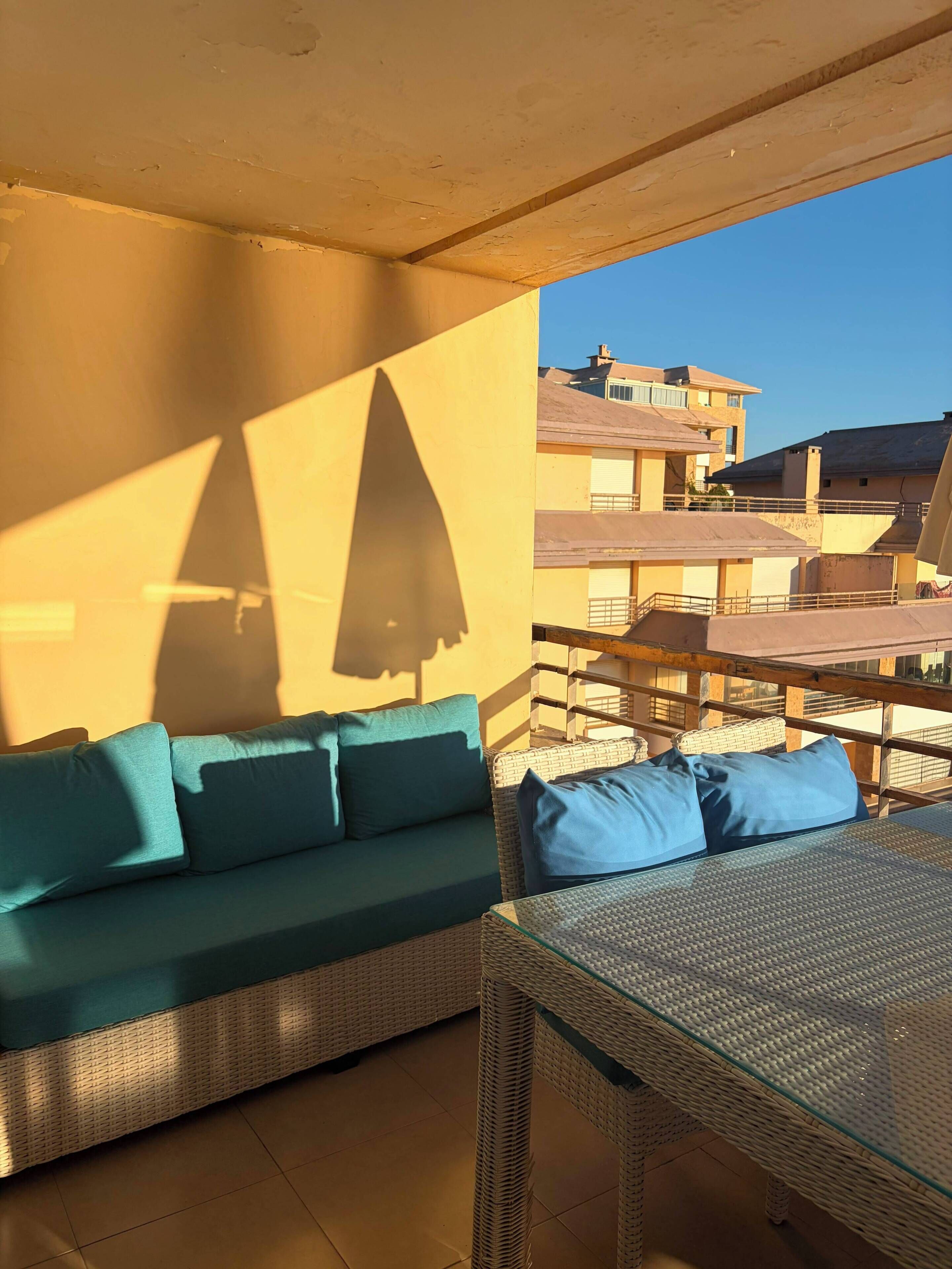 A balcony with a wicker sofa and a glass-top table, overlooking a residential area.