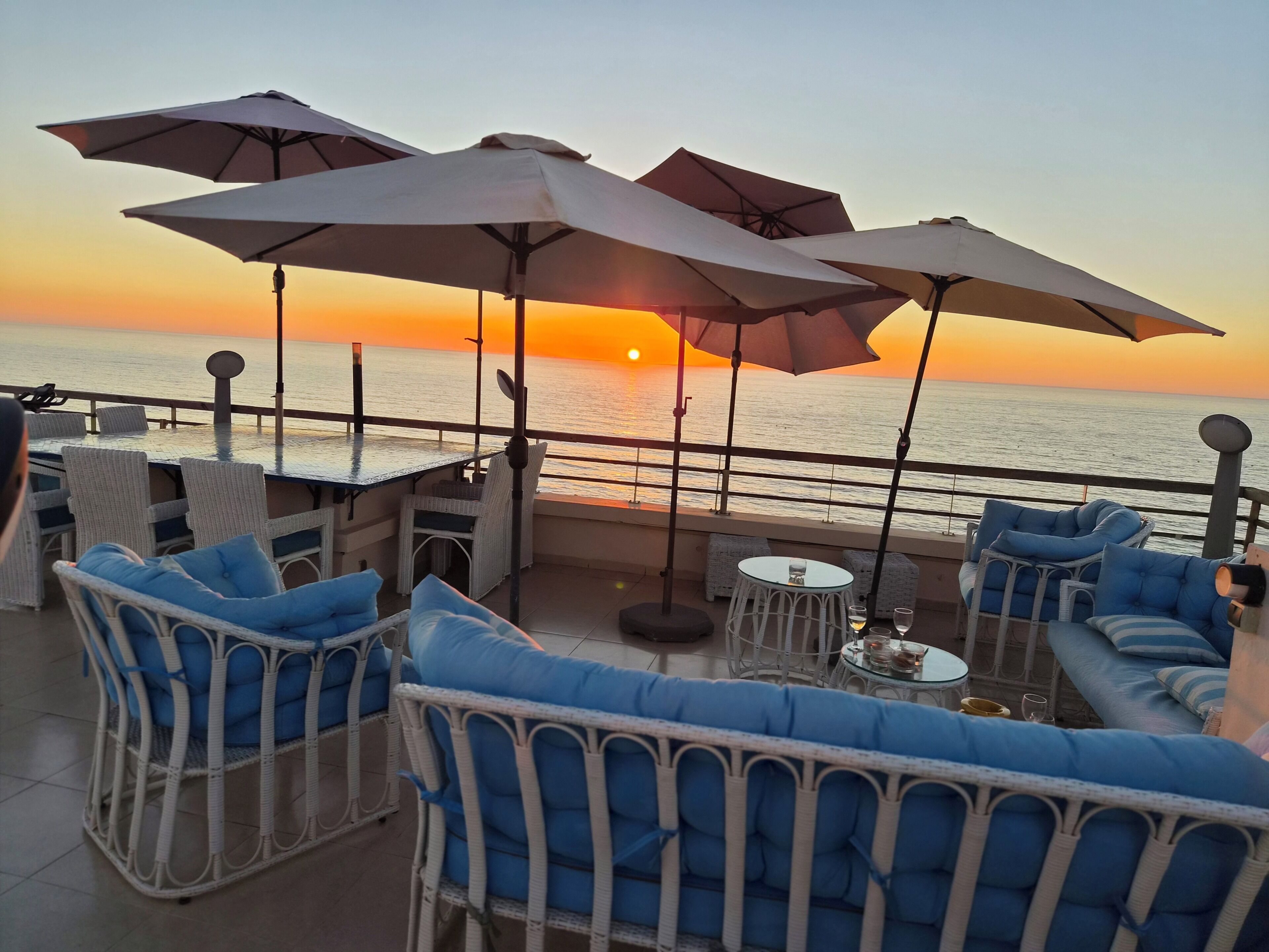 Een terras met zitplaatsen buiten, parasols en uitzicht op de ondergaande zon over het water.
