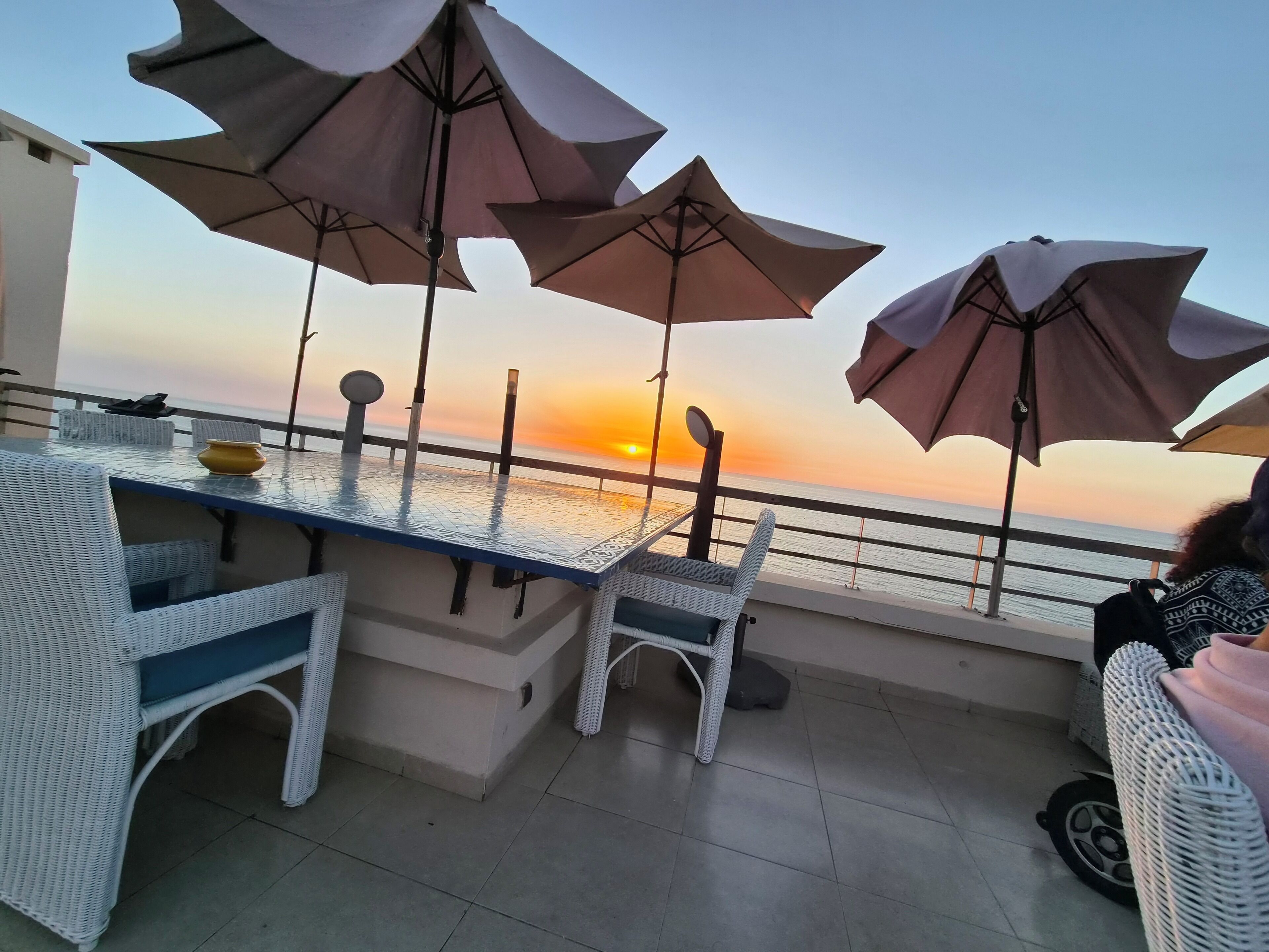 Une terrasse avec une table et des chaises, des parasols et une vue sur le coucher de soleil sur la mer.