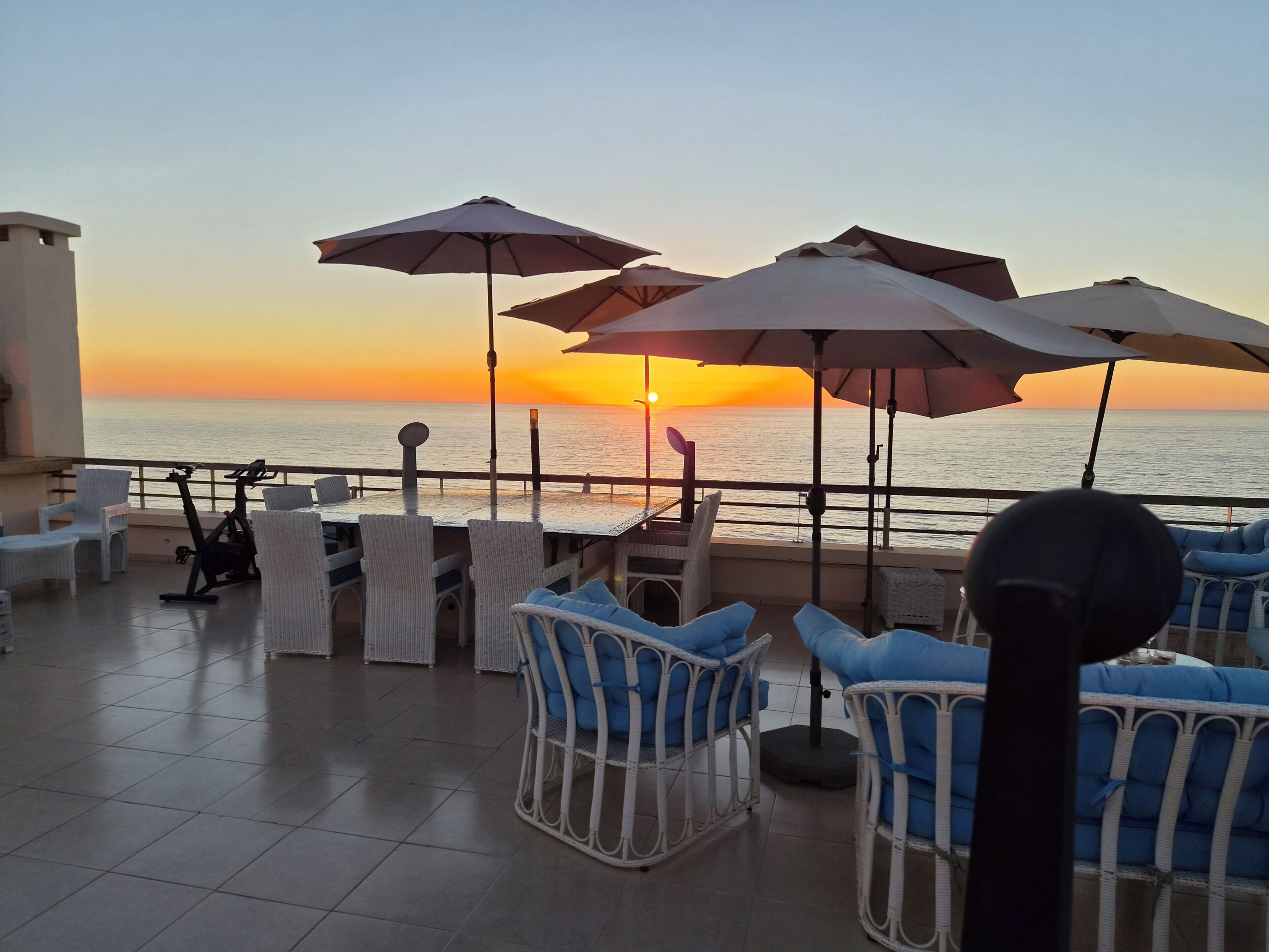Eine Terrasse mit Sitzgelegenheiten im Freien, einem Tisch und Sonnenschirmen mit Blick auf einen Sonnenuntergang über dem Meer.