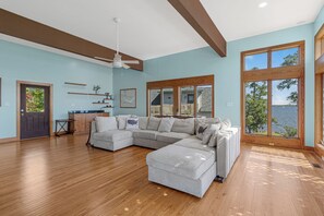 A spacious living room with a large sectional sofa, wooden flooring, and a view of the outdoors through large windows. - Secluded Riverfront Estate with 180 Degree views-Hot Tub, Fire Pit, Kayaks (New Bern)