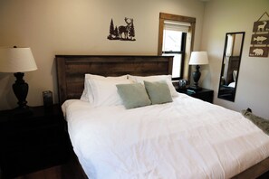 A neatly made bed with white bedding and two pillows, a wooden headboard, bedside lamps, a window with blinds, and wall decorations including a deer and a fish sign. - Rustic Retreat | Fall Lake Retreat | Fire Pit, Kayaks & Paddleboards (New Lisbon)