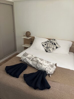 Ein ordentlich bezogenes Bett mit weißen Leinen und einem Kissen mit Blumenmuster, ein Nachttisch mit einer dekorativen Vase sowie ein Paar dunkelblaue Handtücher auf dem Boden.
