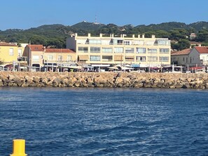 A coastal town with a stone breakwater, buildings with balconies, and a hill with a tower in the background. - At Germaine: Air-Conditioned Apartment with Ocean View (Six-Fours-les-Plages)