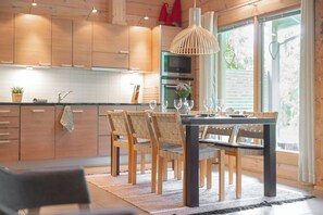 A modern kitchen with wooden cabinets, a dining table set for four, and a large window with a view. - Villa Lumikenkä – Cozy Retreat & Private Sauna (Kuusamo)