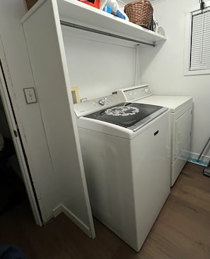 A compact laundry room with a washer and dryer, a shelf with a basket, and a window with blinds.