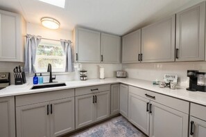 A kitchen with white cabinets, a sink, a window with curtains, a coffee maker, and a sign that says 'Coffee Time'. - Saltwater Heated Pool | Kayaks |Golf Cart | Firepit | Beach (Palm Coast)