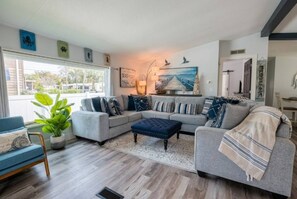 A modern living room with a sectional sofa, a blue armchair, and a coffee table. There is a large window providing natural light and a view of the outdoors. - Saltwater Heated Pool | Kayaks |Golf Cart | Firepit | Beach (Palm Coast)