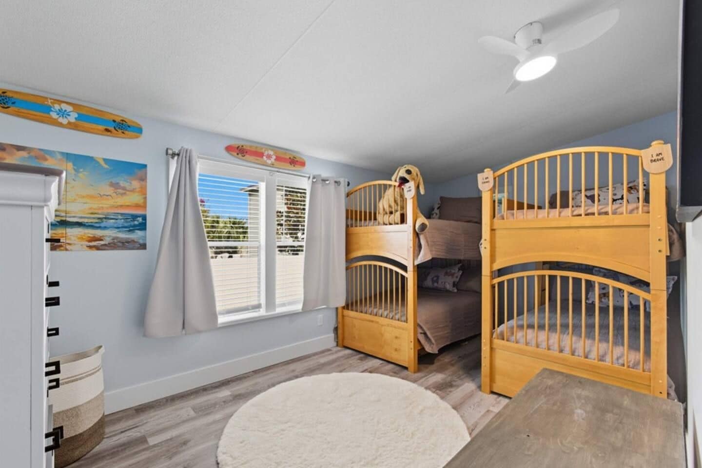 A room with two bunk beds, a ceiling fan, a window with curtains, a painting, and a small rug.