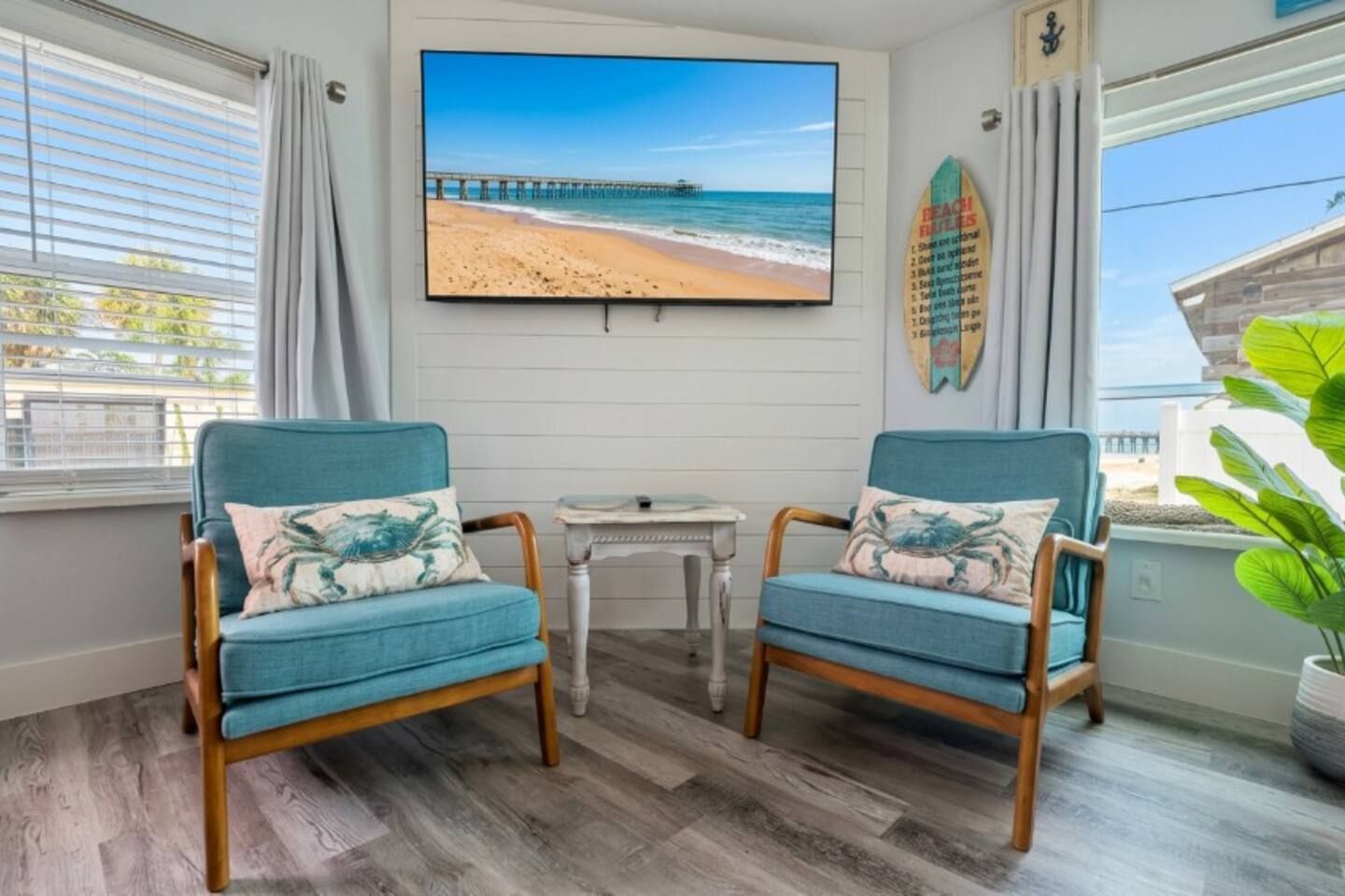 A room with two blue armchairs, a small table, a television displaying a beach scene, and a surfboard on the wall.