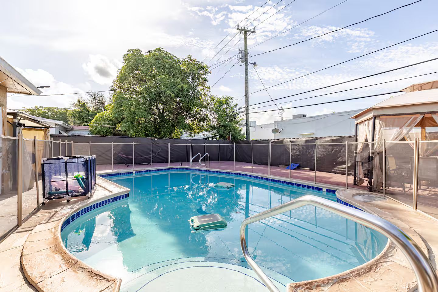 Una piscina con un deck cercado, rodeada por una zona cubierta y un árbol.