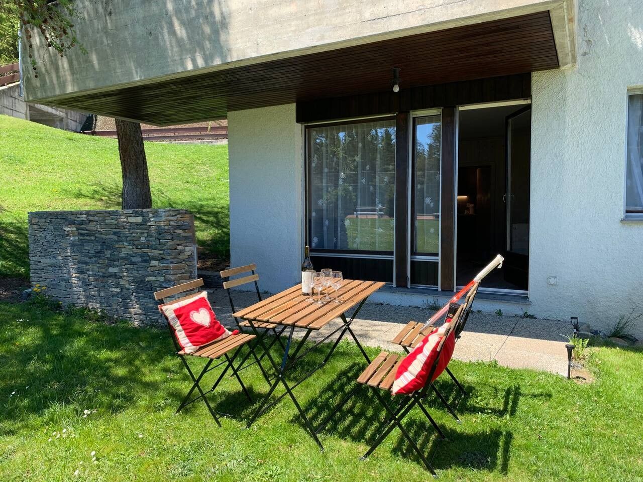 An outdoor seating area with a table and chairs, a stone wall, and a building entrance with glass doors.