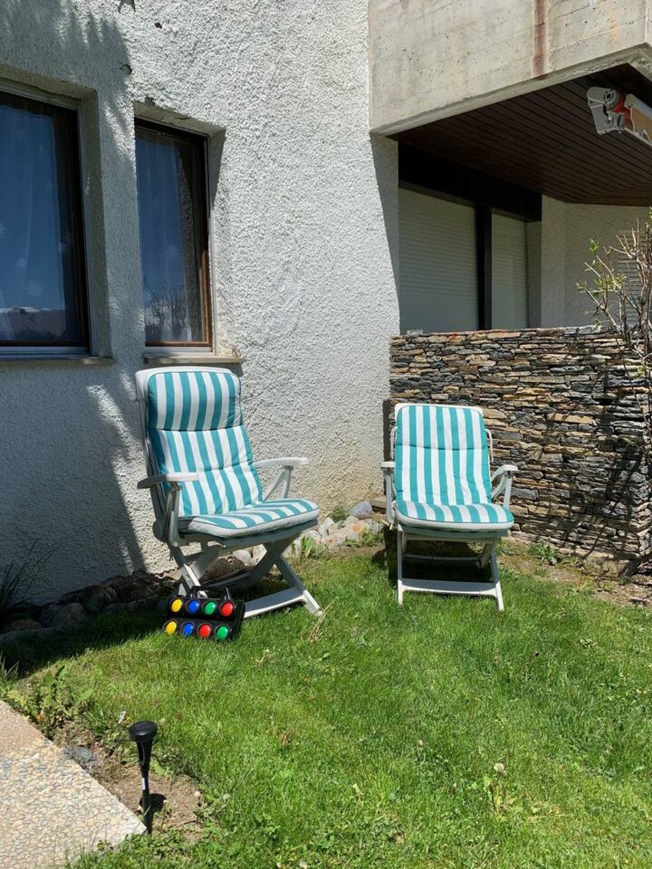 Deux fauteuils de jardin à rayures sont placés à l’extérieur d’un bâtiment doté d’un mur en pierre et d’une fenêtre.