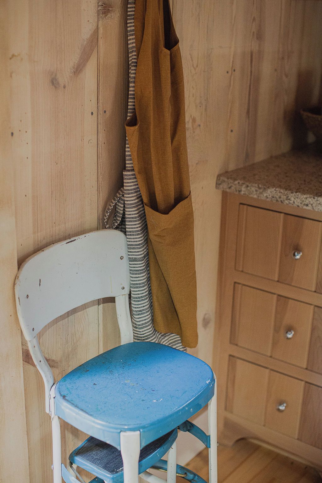 A chair with a blue seat and white backrest, a brown apron hanging on a wooden wall, and a wooden cabinet with drawers.