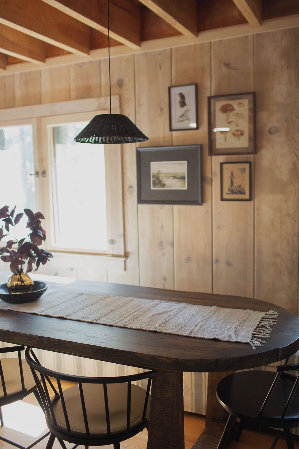 A wooden dining table with a woven placemat, black chairs, and a hanging pendant light.