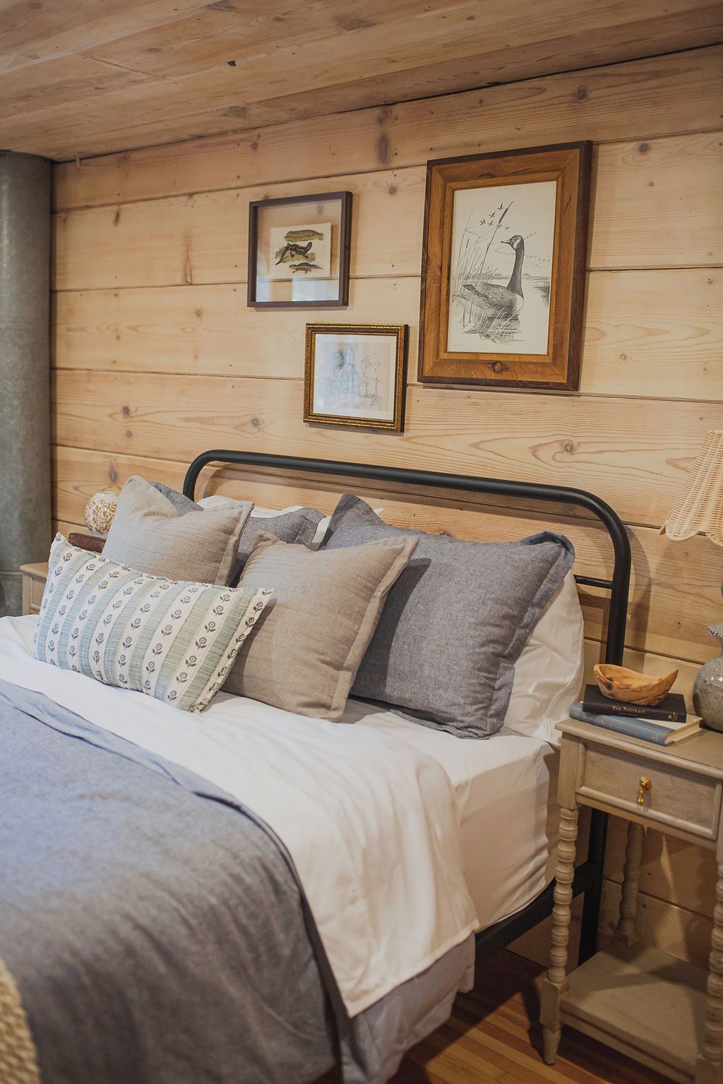 A wooden-paneled room with a bed, pillows, a nightstand, and framed artwork on the wall.