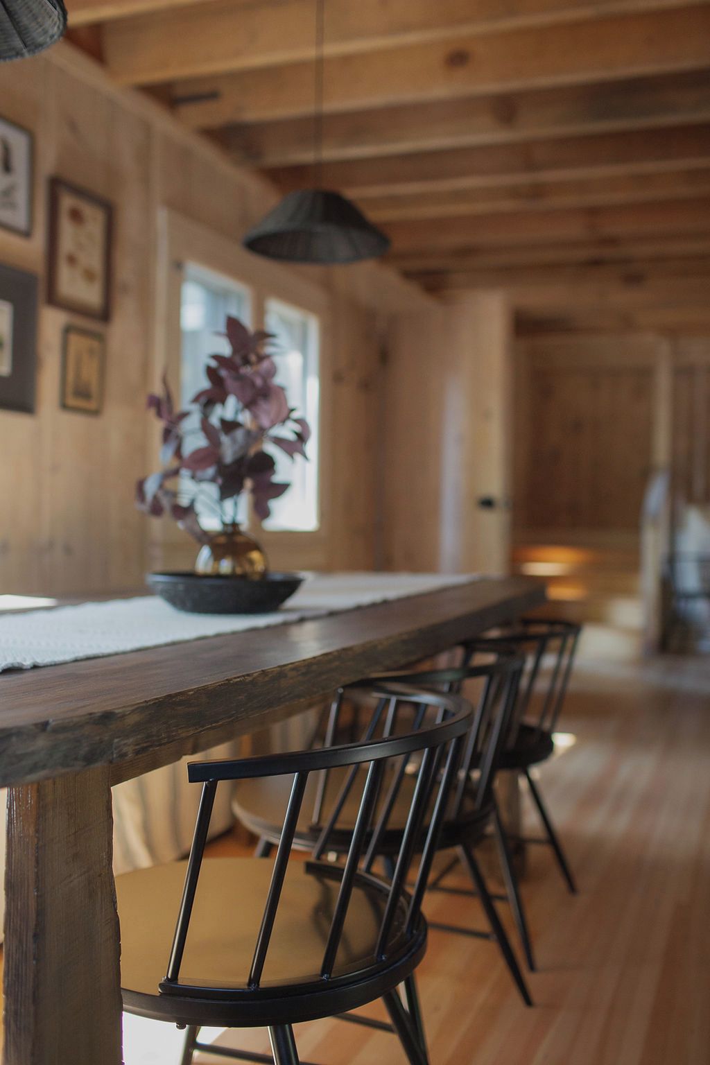 A wooden interior with a long table and chairs, a hanging light fixture, and a window with a plant.