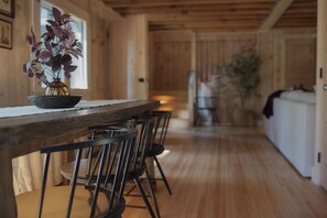Un interior de madera con una mesa larga y sillas, un jarrón con flores moradas y vistas a otra habitación.